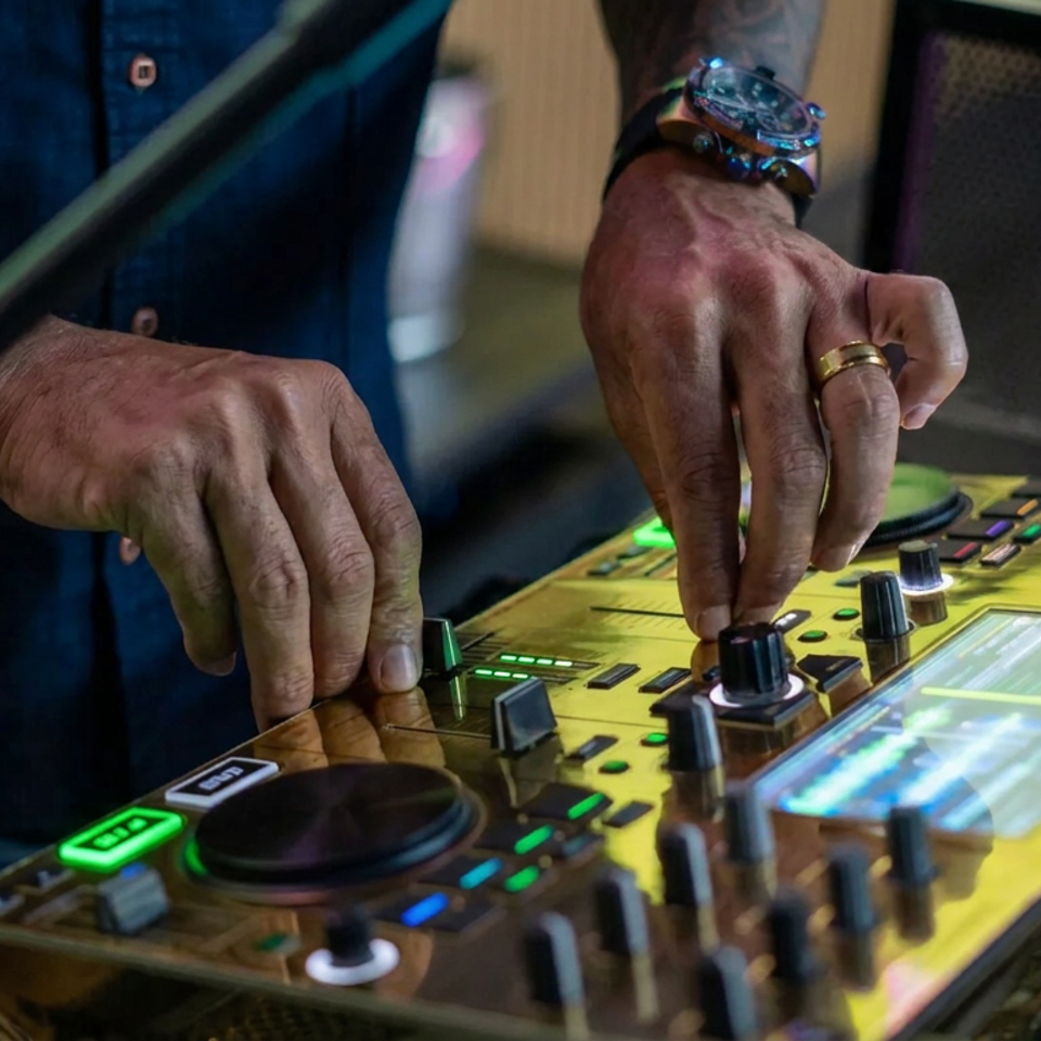 Tattooed DJ adjusts knobs on a brightly lit Pioneer DJ mixer during a live performance.