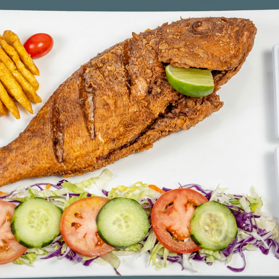 Crispy whole fried fish served with fries, tomato-cucumber salad, lime, and two dipping sauces on a white plate.
