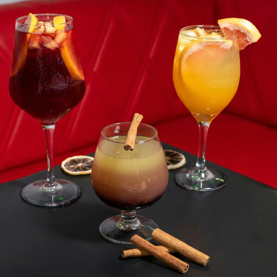 Three colorful cocktails sit on a black table against a red booth, garnished with citrus slices and cinnamon sticks.